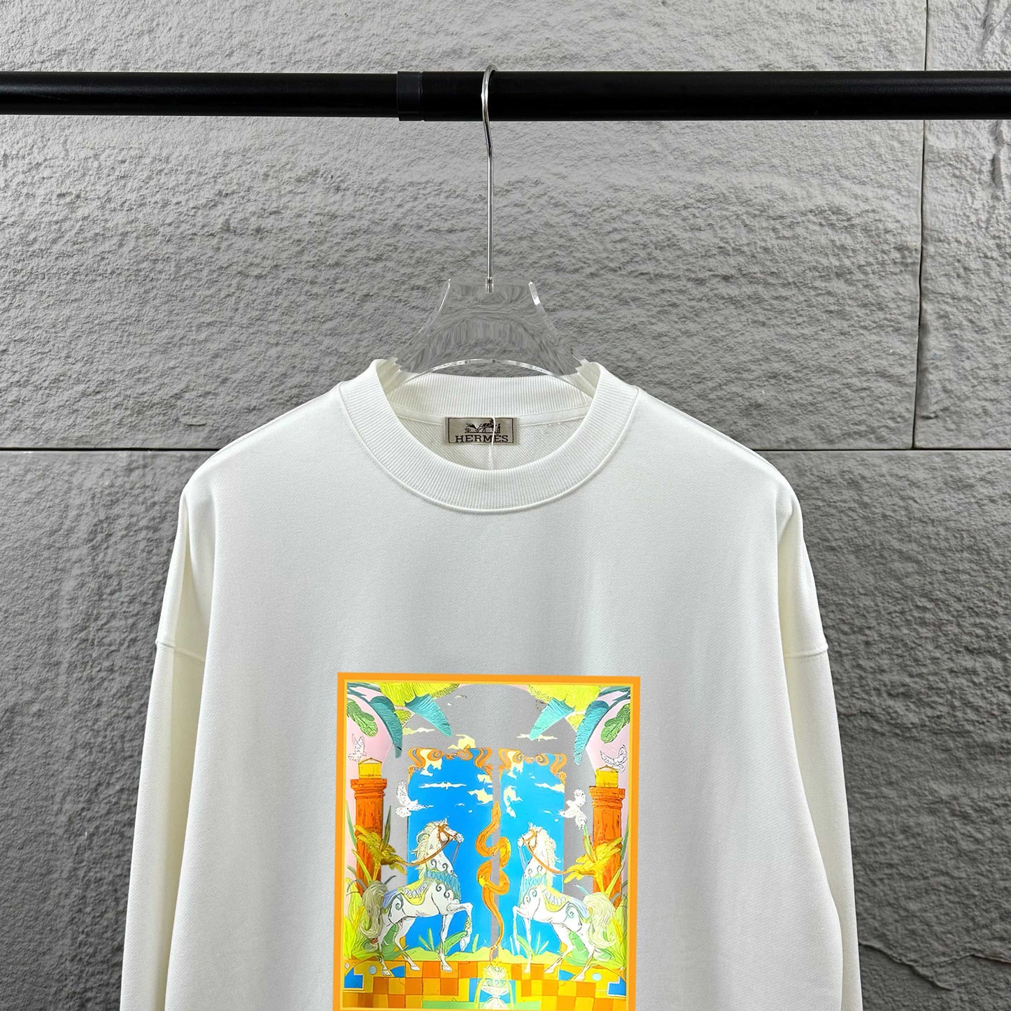 White sweatshirt with colorful graphic design on a hanger against a gray wall.