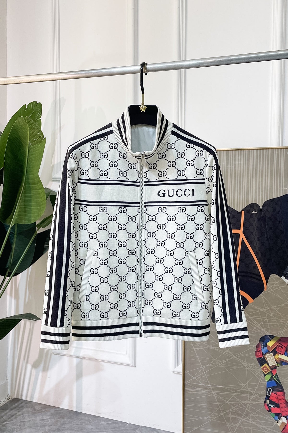 A  zip-up jacket featuring the brand's signature GG monogram pattern in black and white, hanging on a black hanger. The jacket has a white base with black monogram print, black and white striped ribbed collar, cuffs, and hem, and a black and white horizontal stripe across the chest with 'printed in black.