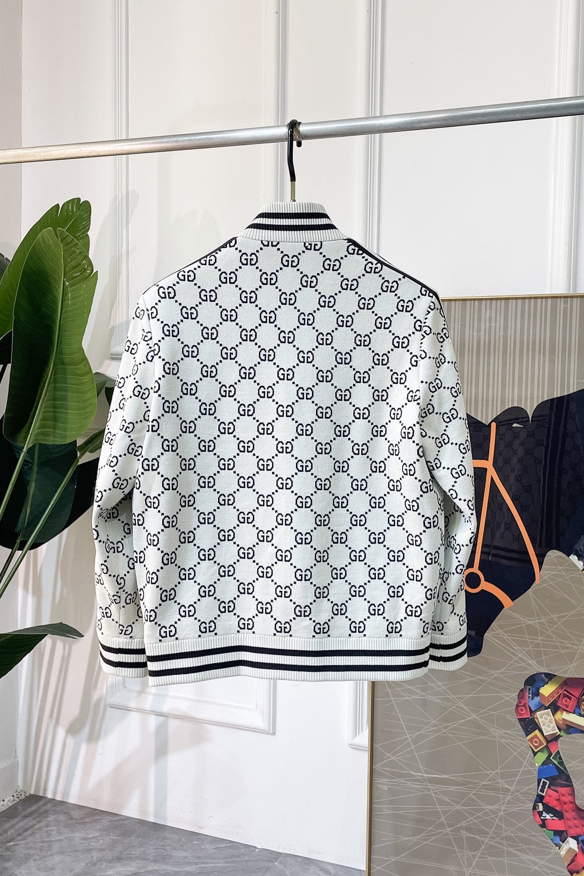 A white bomber jacket with a black  GG monogram pattern, hanging on a metal rack in a boutique setting.