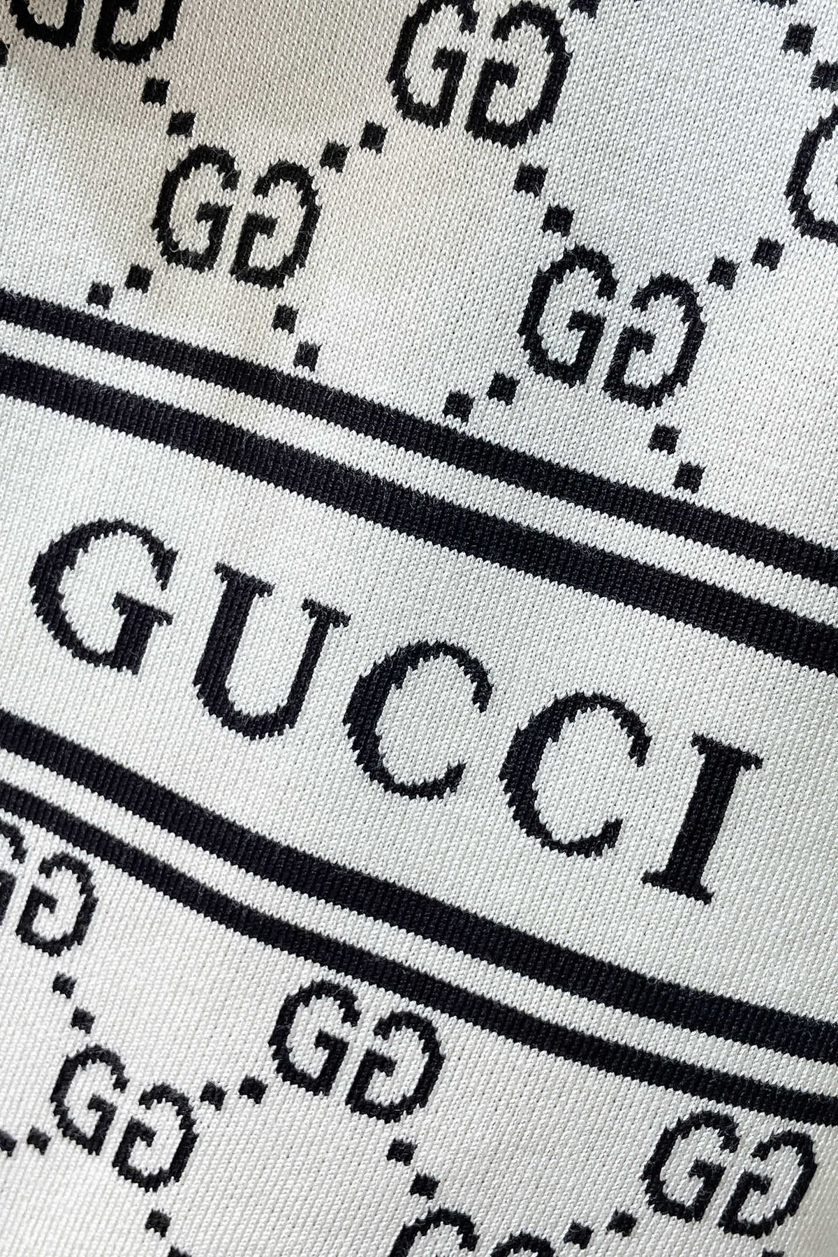 Close-up of a textile featuring the brand name in large black letters between two horizontal black stripes, with a repeating GG monogram pattern on a light background.